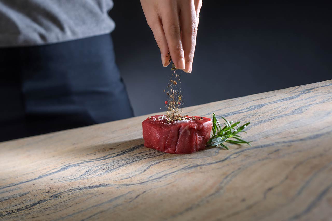 Küchenarbeitsplatte aus Naturstein, auf der ein Steak gewürzt wird.