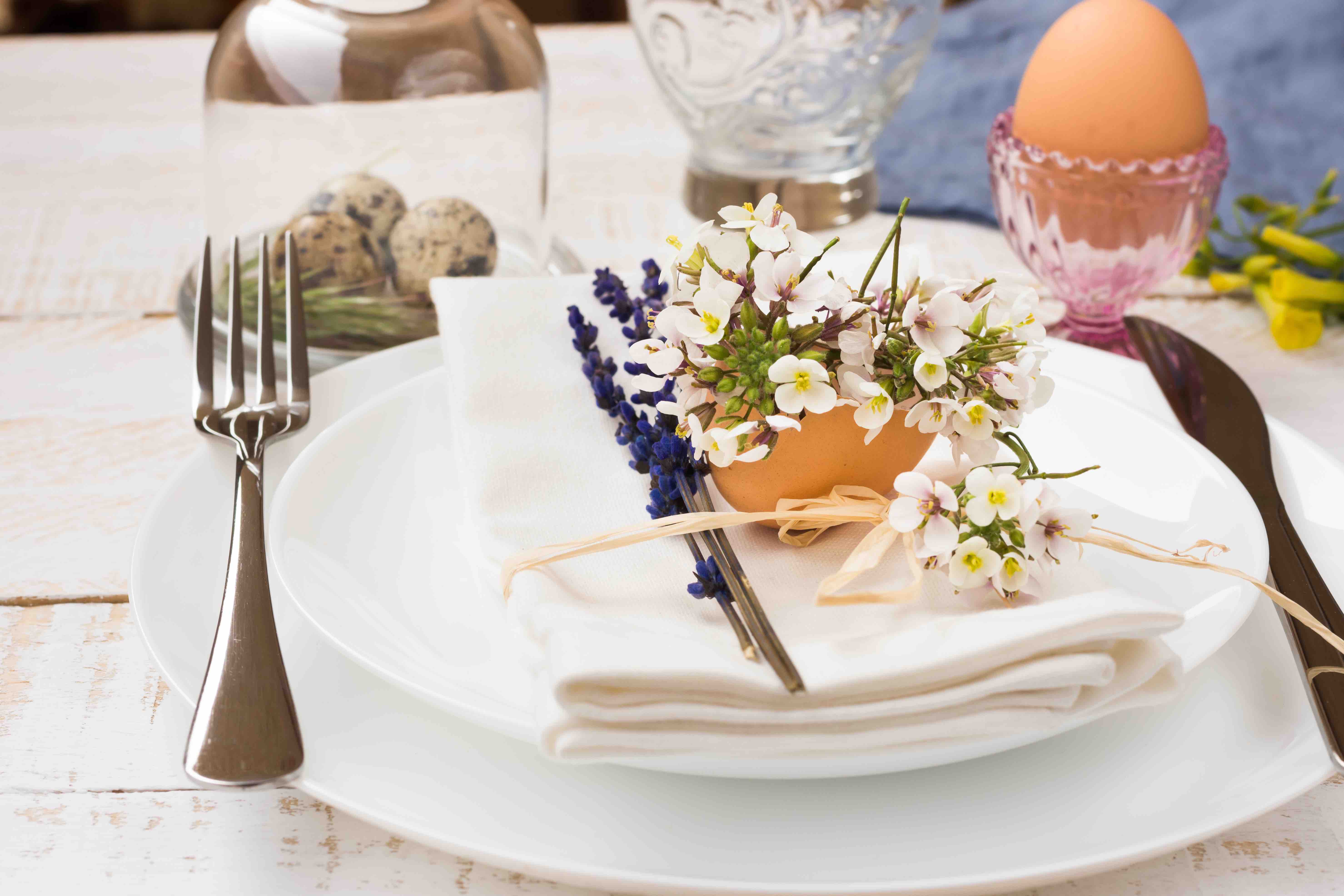 Weißer Holztisch mit weißem Geschirr, weißer Stoffserviette und Besteck. Auf dem Gedeck liegt eine Deko mit Eierschale, Lavendel und weißen Blumen. Auf dem Tisch stehen ein rosafarbener Glas-Eibecher mit gekochtem Ei darin sowie eine Glasglocke mit drei Wachteleiern darunter.