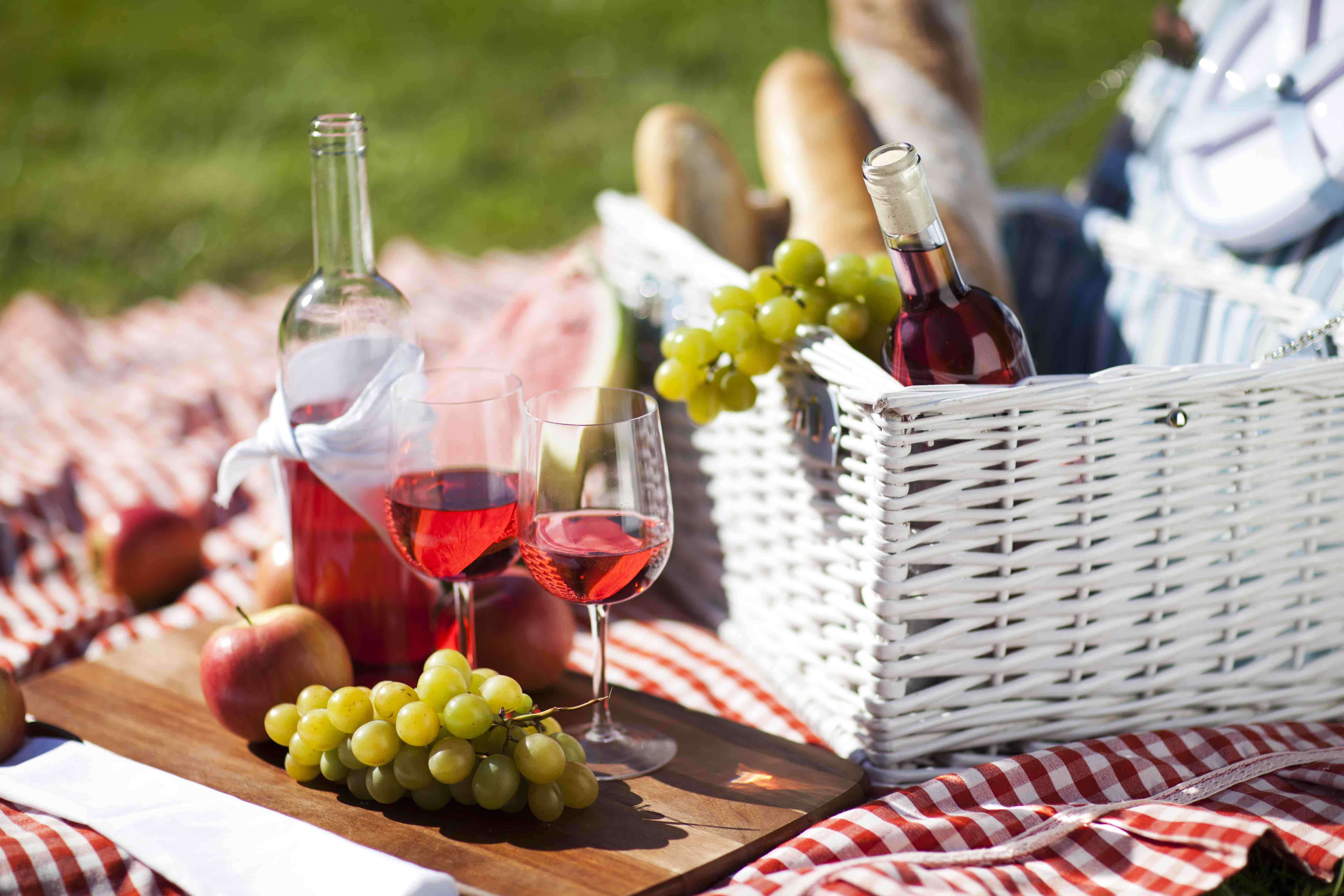 Rot-weiß karierte Picknickdecke auf der Wiese mit weißem Picknick-Korb, Baguettes, Weintrauben und Wein.