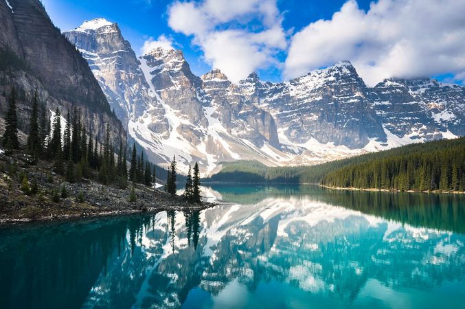 Blauer See mit Wald drumherum und schneebedeckten Bergen im Hintergrund in Kanada.