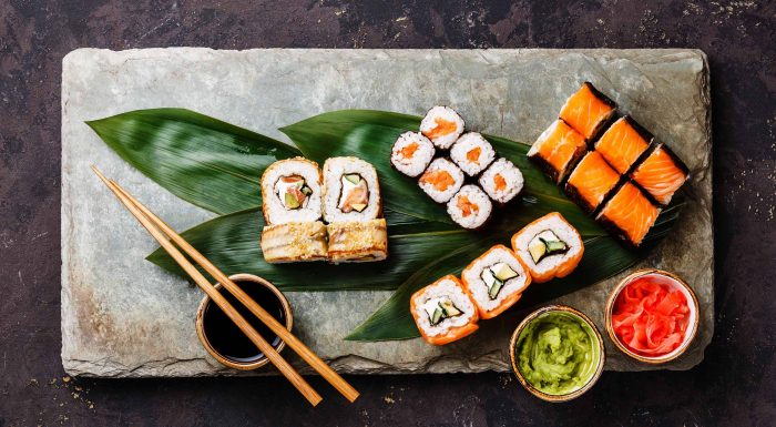 Sushi angerichtet auf einer Steinplatte mit Essstäbchen, Blättern und Schälchen mit Sojasauce, Wasabi und eingelegtem Ingwer.