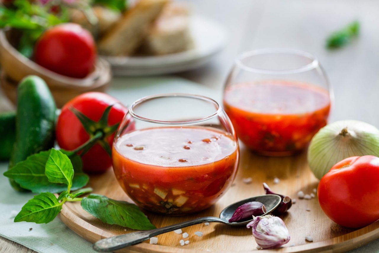 Gazpacho in Gläsern auf einem Holztablett, mit Tomaten und Basilikum im Hintergrund.