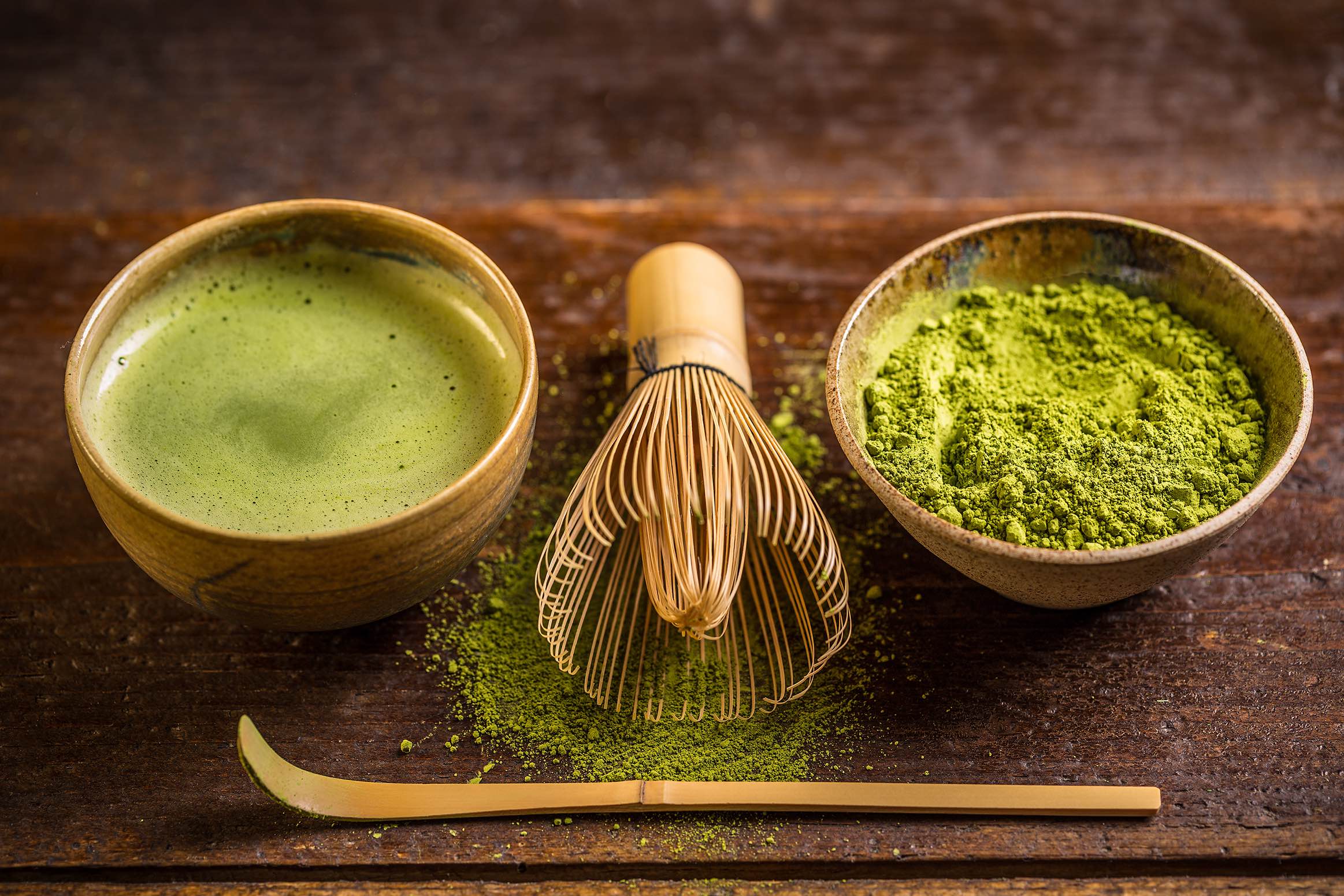 aufgeschäumter Matcha-Tee in einer Bambustasse, daneben ein kleiner Bambusbesen und eine schale mit Match-Teepulver.